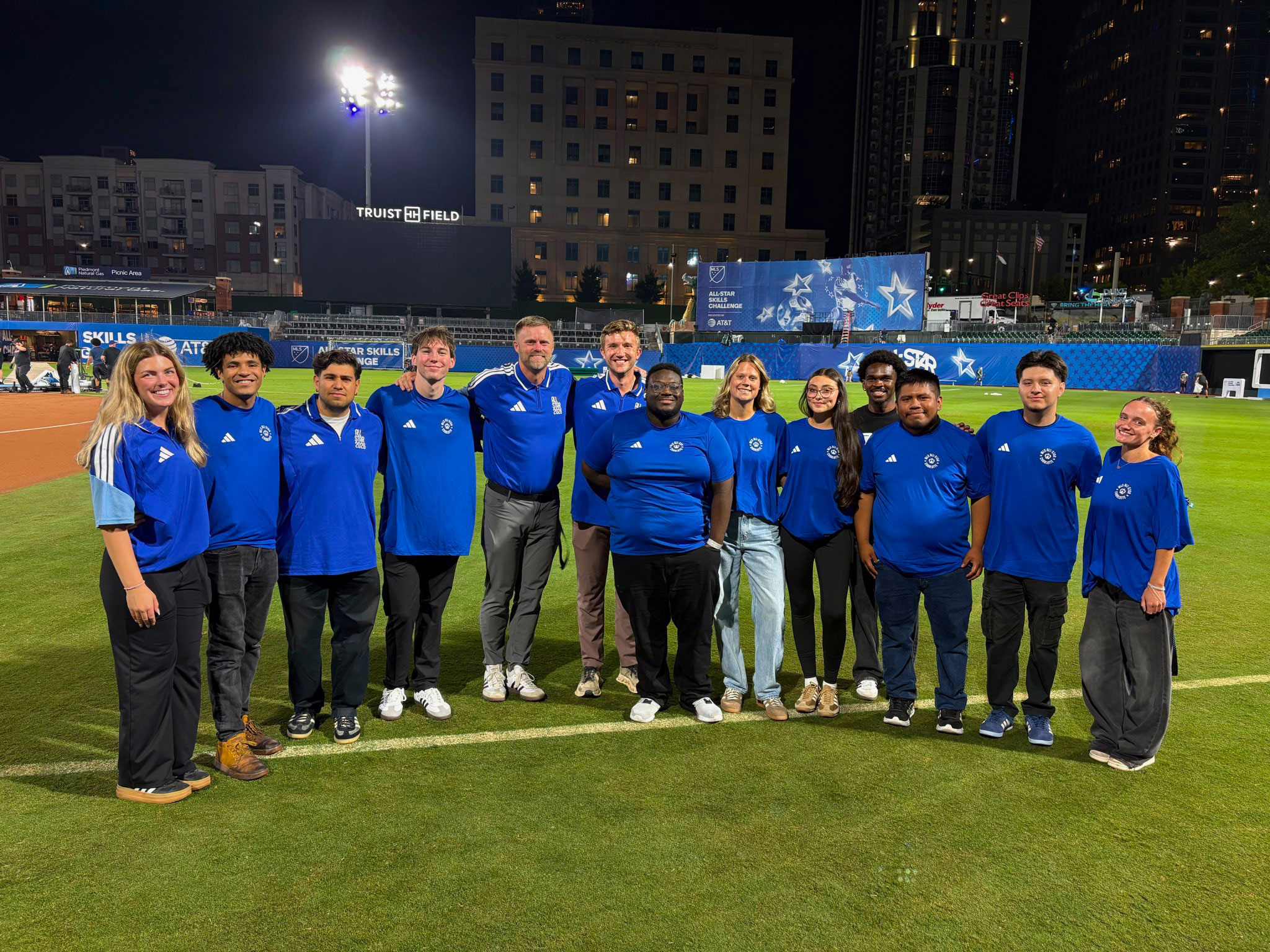 A group of people with blue shirts and black pants stand on an the athletic field at Bank of America