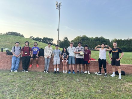 A group of Elon Academy Scholars line up for a picture with their soccer instructor in front of Elon University's Intramural fields.