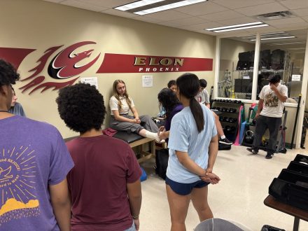 Two instructors showcase ankle taping techniques to a group of students in an athletic training style room. One instructor sits on the athletic training bed while the other tapes her ankle. 