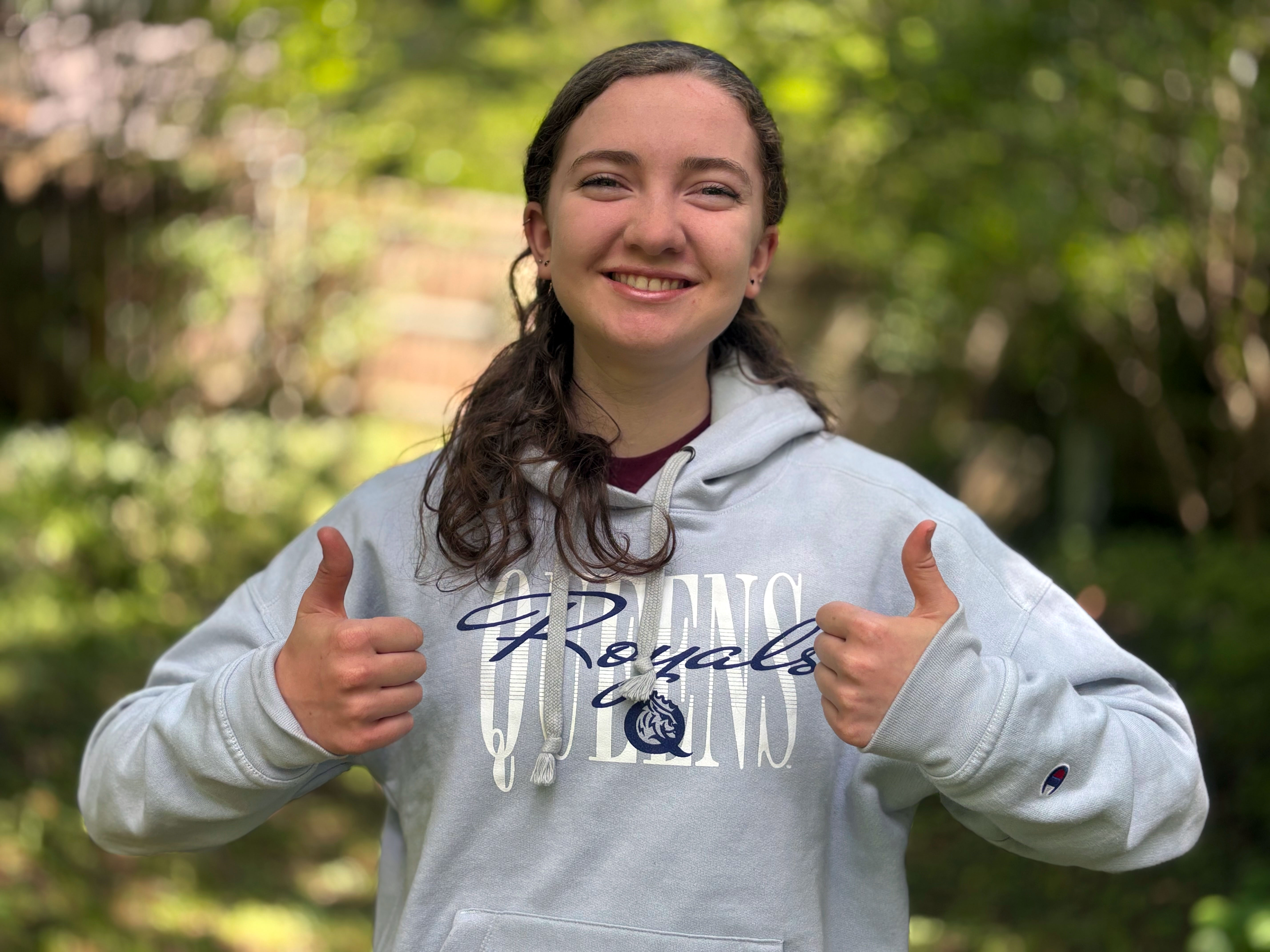 Girl wearing a gray Queens Royals sweatshit gives two thumbs up on a nature trail.