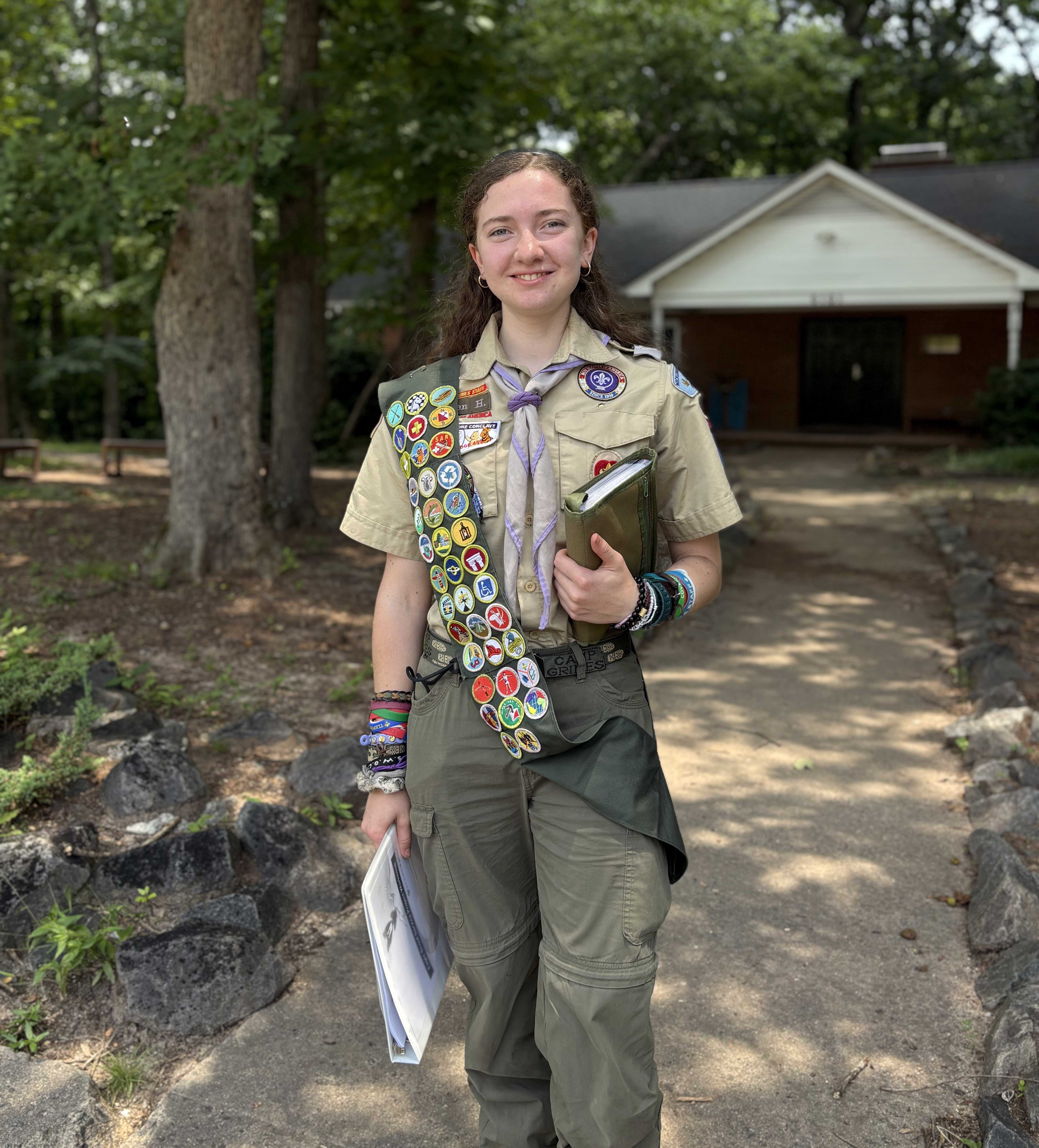 Girl dressed in Scouting Ameria apparel with sash of badges stands in a camp ground.