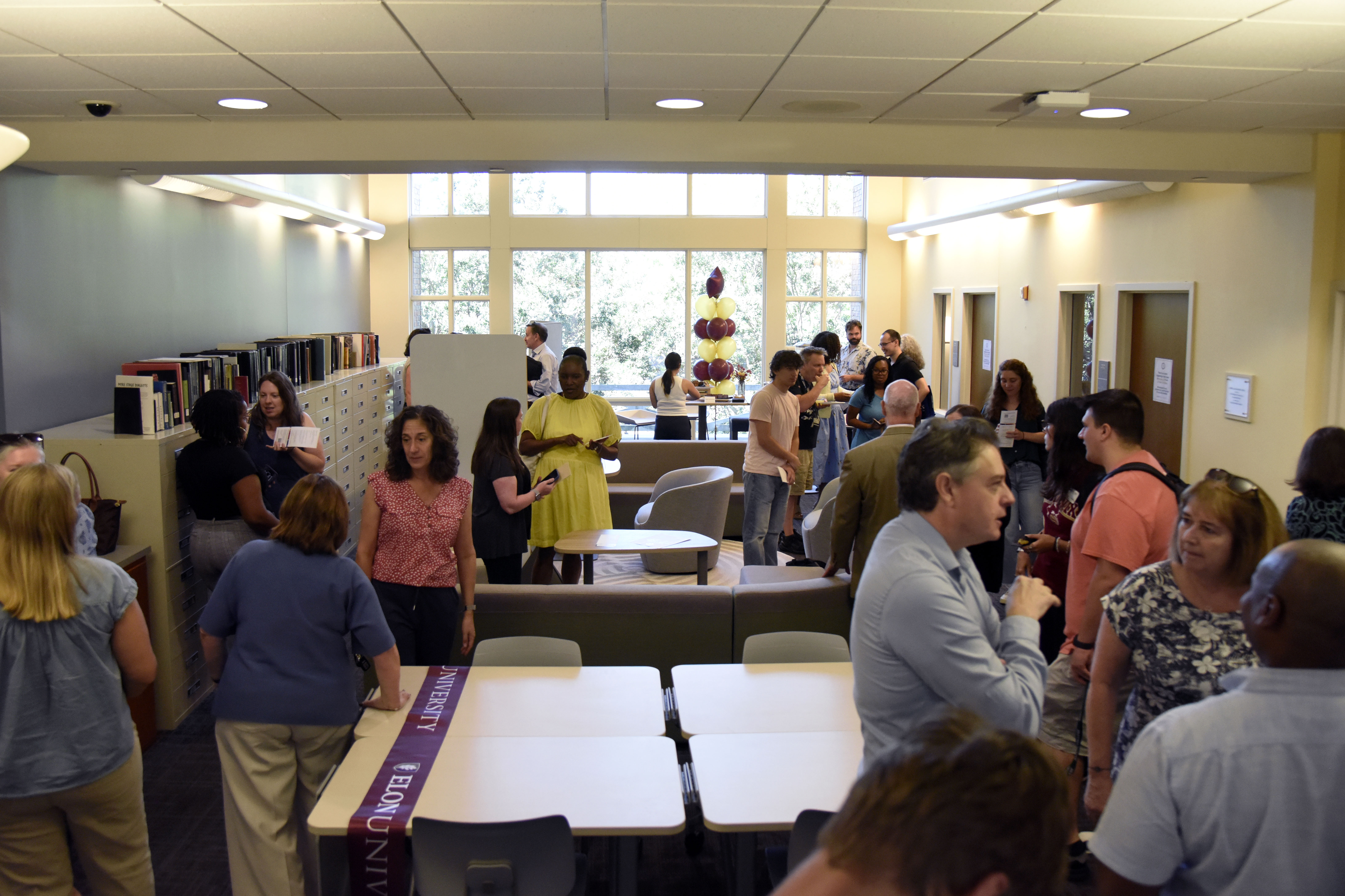 A photo of Elon University faculty, staff and students touring redesigned study space on the second floor of Belk Library with a large window against the far wall, and tables and chairs in the room.