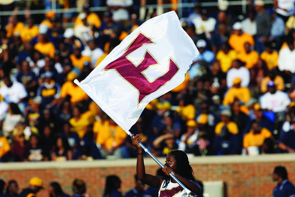An Elon cheerleader waves a large flag featuring the Elon “E” monogram in front of a packed football stadium.