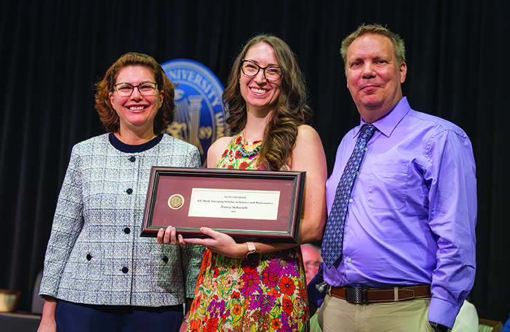 A woman holds a framed Elon University award while posing with two people during a ceremony.