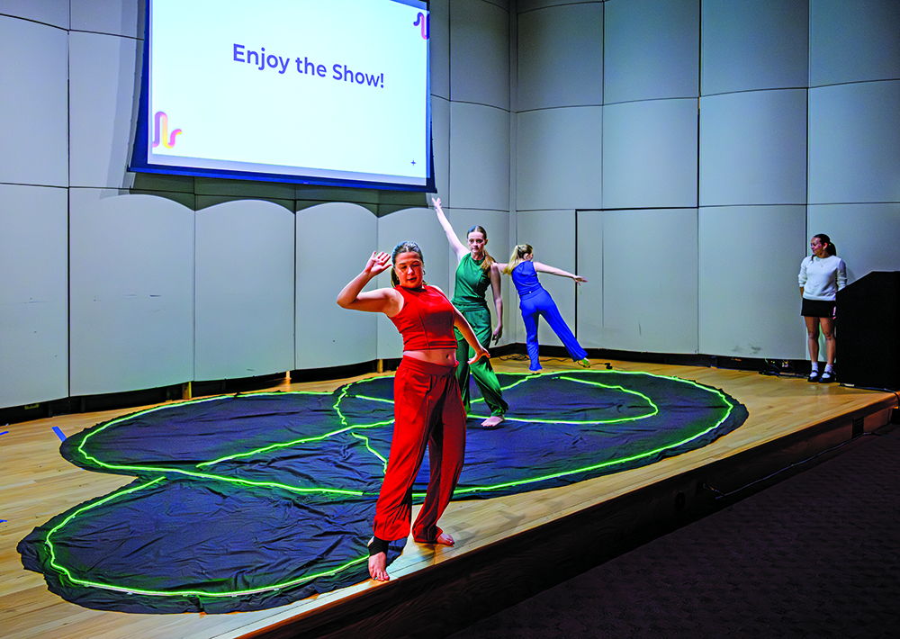 Three student dancers perform in red, green and blue costumes onstage during a presentation.