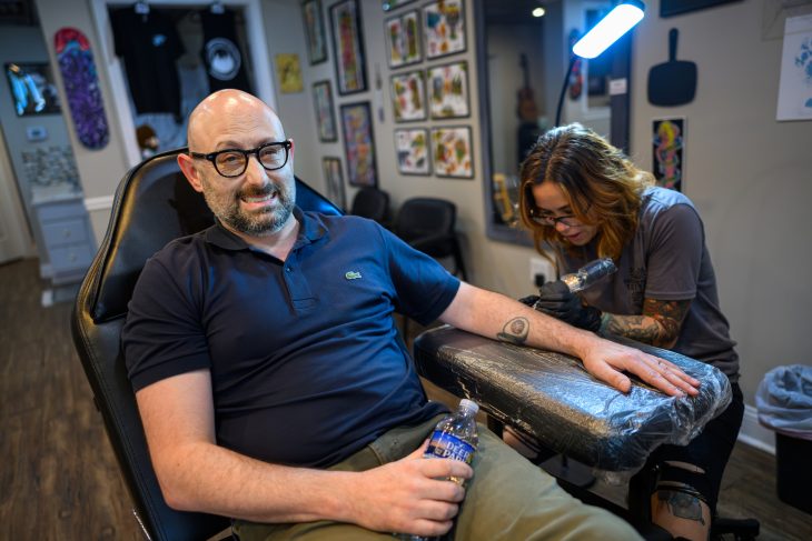 A man makes a face of discomfort as a tattoo artist puts the needle on his left arm.