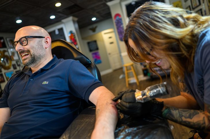 A tattoo artist leaned over a man's forearm as he laughs in a chair.