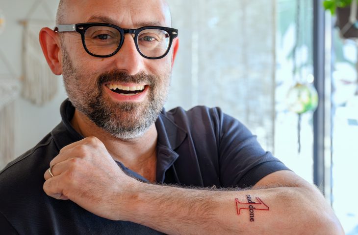 Dean Zak Kramer shows off the tattoo of his 1 and Done slogan, smiling