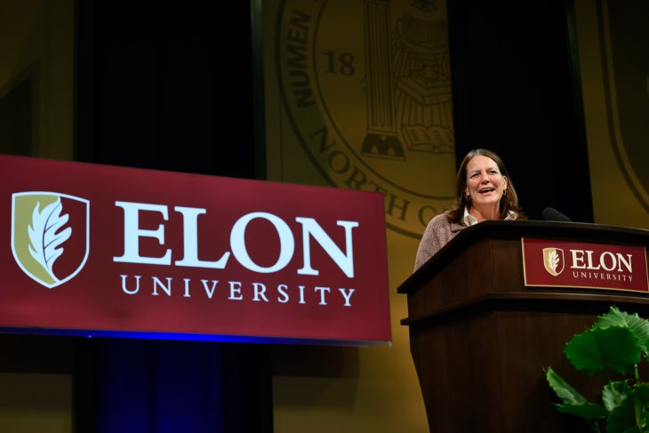 A woman speaks from an Elon University podium onstage, smiling as she addresses the audience during Fall Convocation.