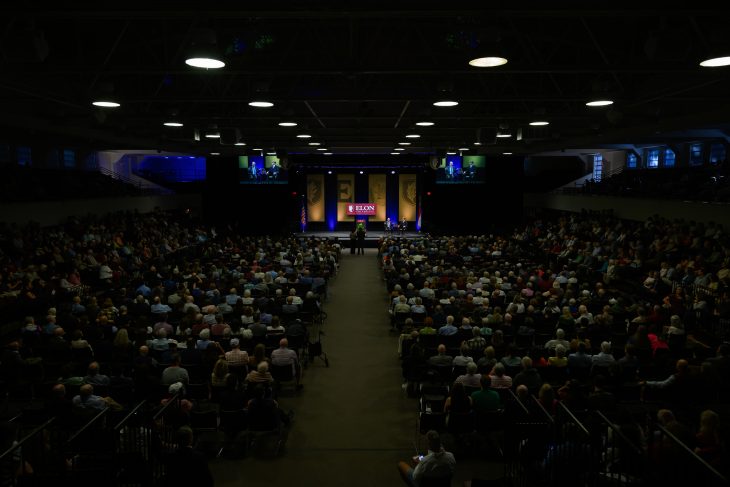 A large audience fills Alumni Gym for Elon University’s Fall Convocation, with David Brooks and an Elon student seated onstage.