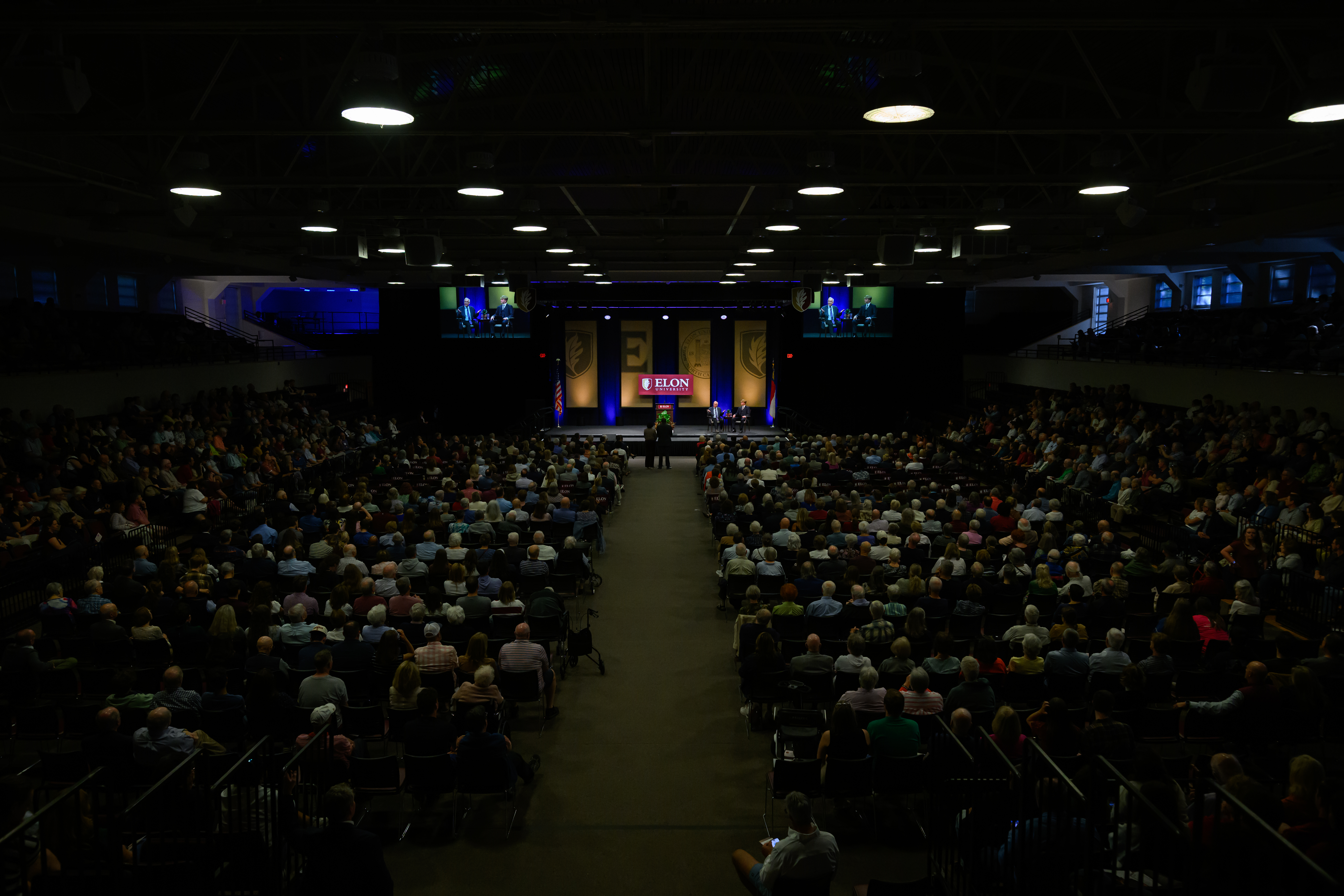 A large audience fills Alumni Gym for Elon University’s Fall Convocation, with David Brooks and an Elon student seated onstage.