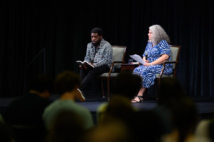     A guest speaker reads from a book onstage while a moderator sits beside him holding a microphone.
