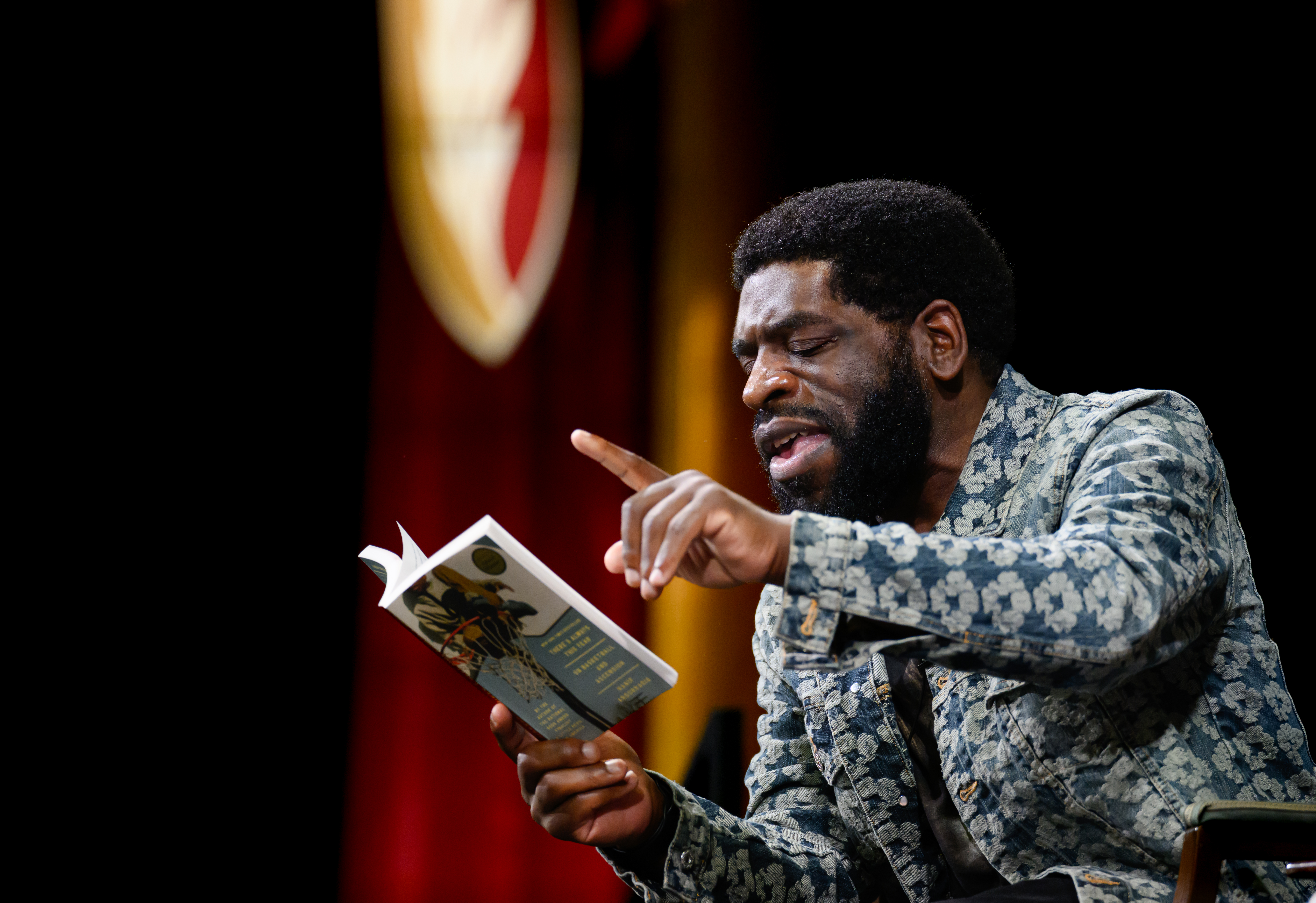 A guest speaker gestures while reading from a book during Elon University’s Common Reading lecture.