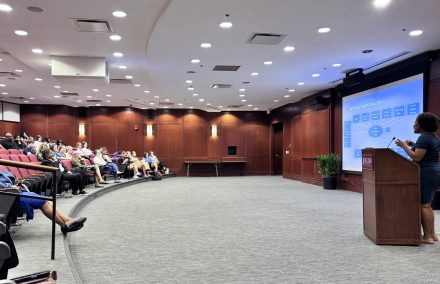 A woman speaks in a lecture hall setting behind an Elon University labeled podium