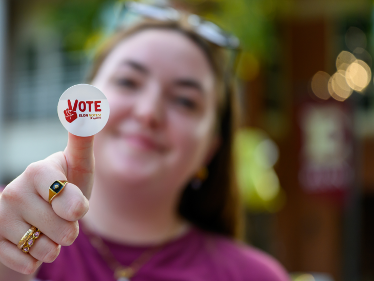 Student smiling and holding up a thumb with a 'Vote, Elon Votes!' sticker at the campus Organization Fair.