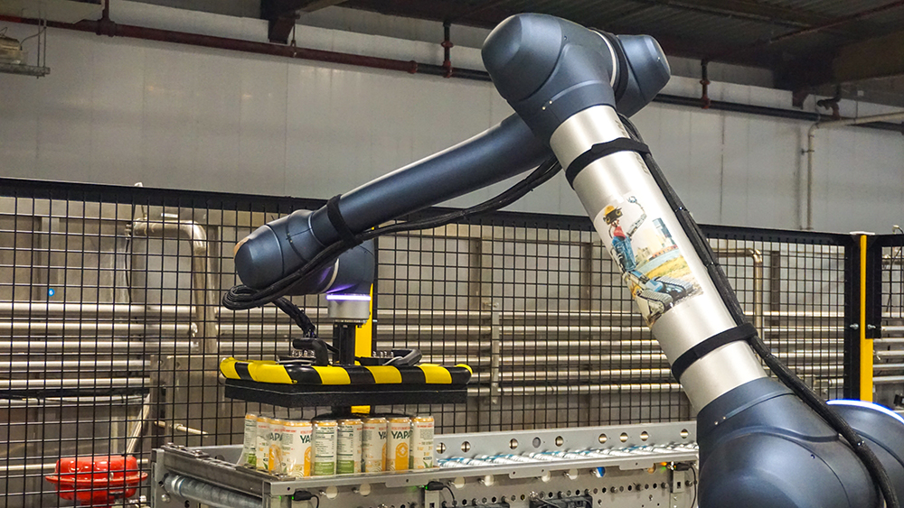 A robotic arm moves cans of YAPA energy drinks along a production line.