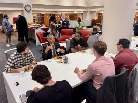 a group of people seated around a large table conversing. People in the background are in small groups talking.