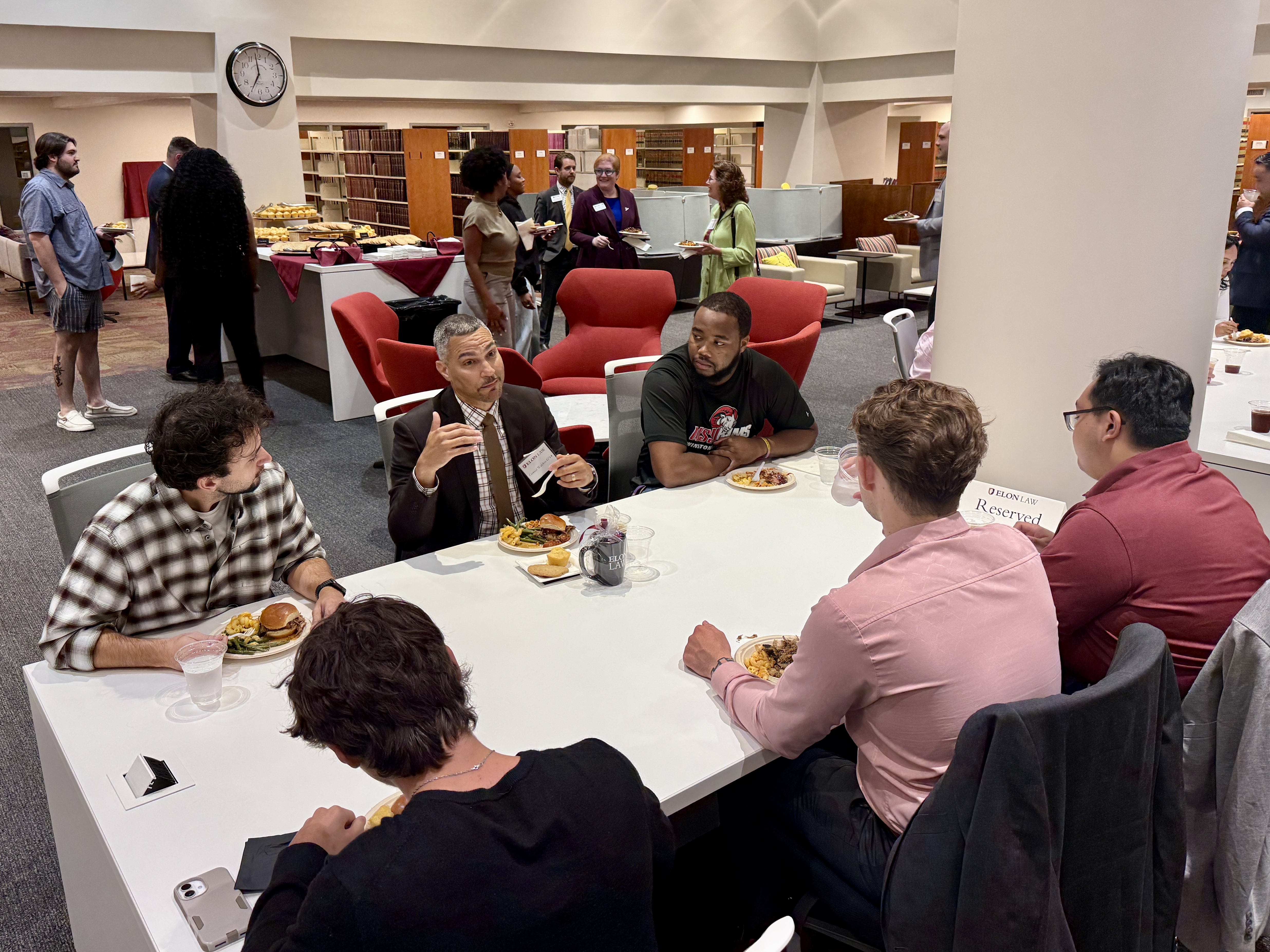 a group of people seated around a large table conversing. People in the background are in small groups talking.