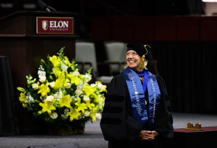 A smiling woman in academic regalia and an Elon University first-generation stole stands onstage during a ceremony.