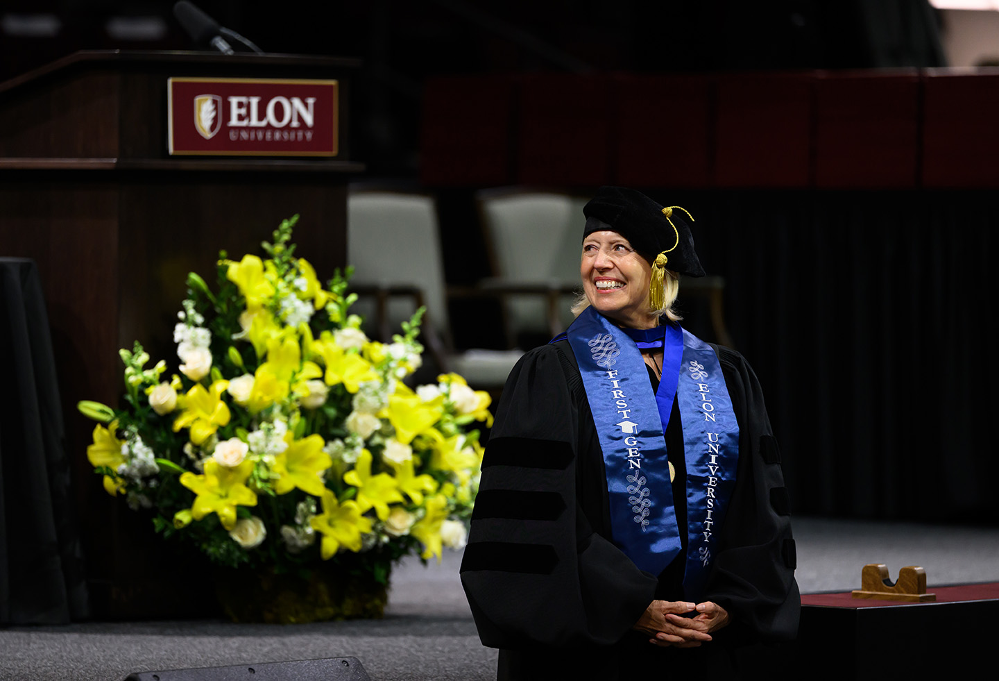 A smiling woman in academic regalia and an Elon University first-generation stole stands onstage during a ceremony.