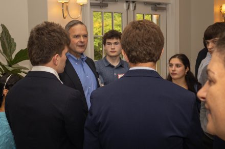 A man speaks with a group of students during a gathering, surrounded by attendees in a crowded room.