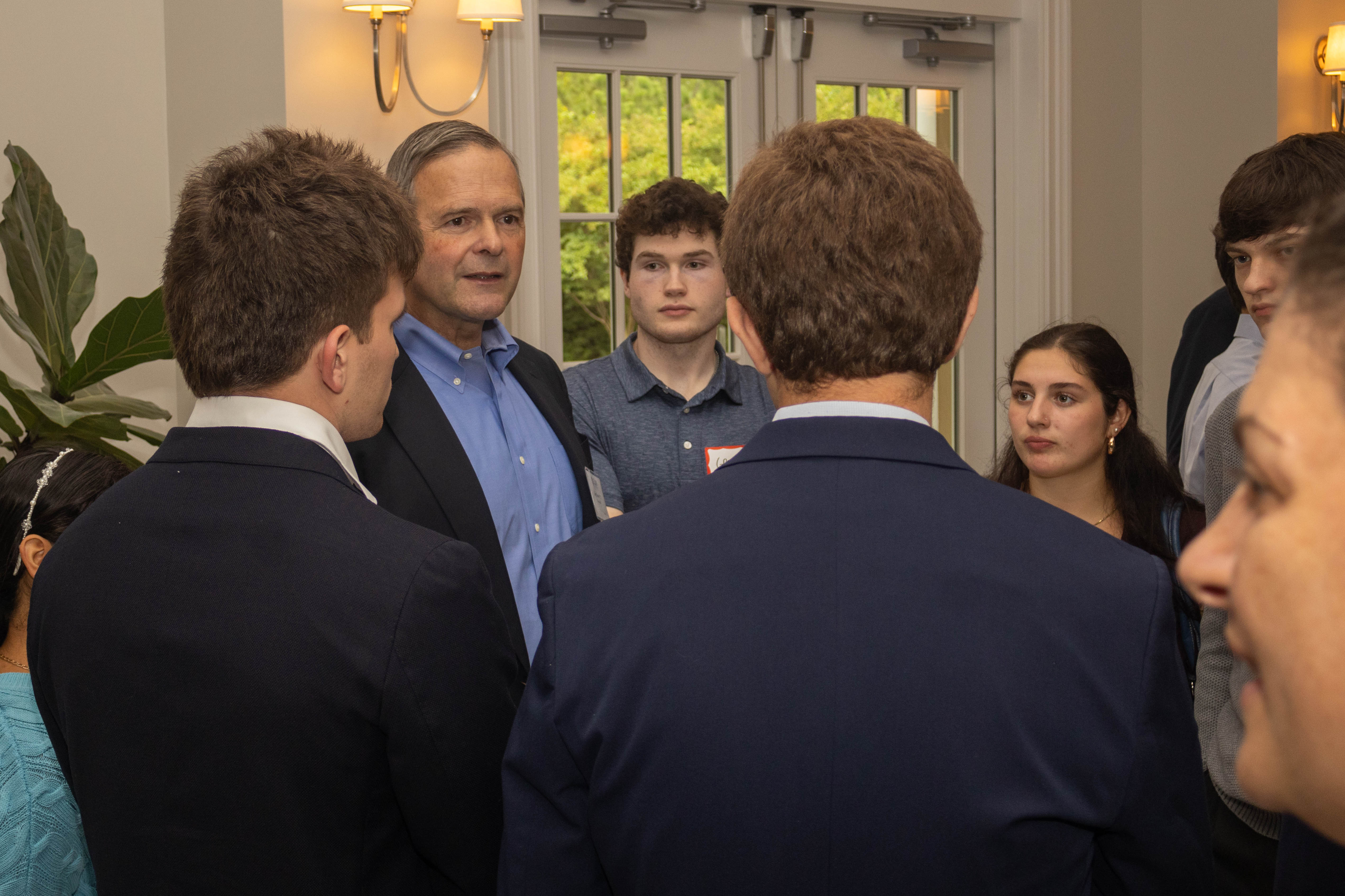 A man speaks with a group of students during a gathering, surrounded by attendees in a crowded room.