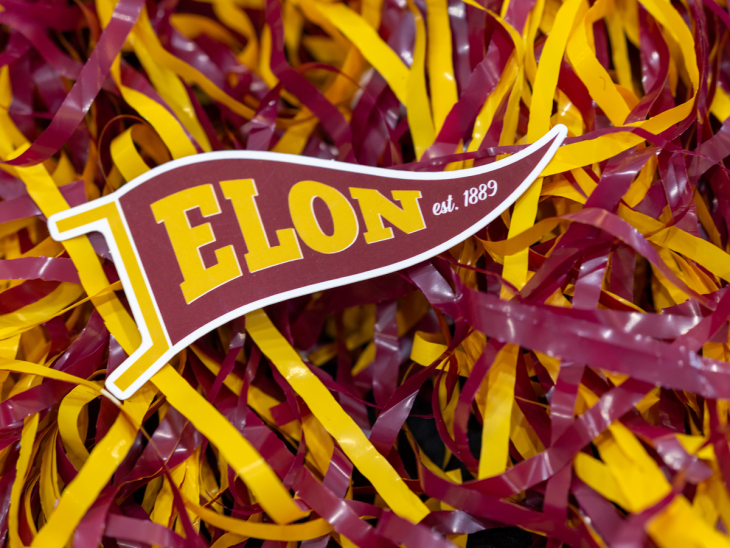 Elon, est. 1889' pennant resting on maroon and gold shredded paper, likely for Homecoming.