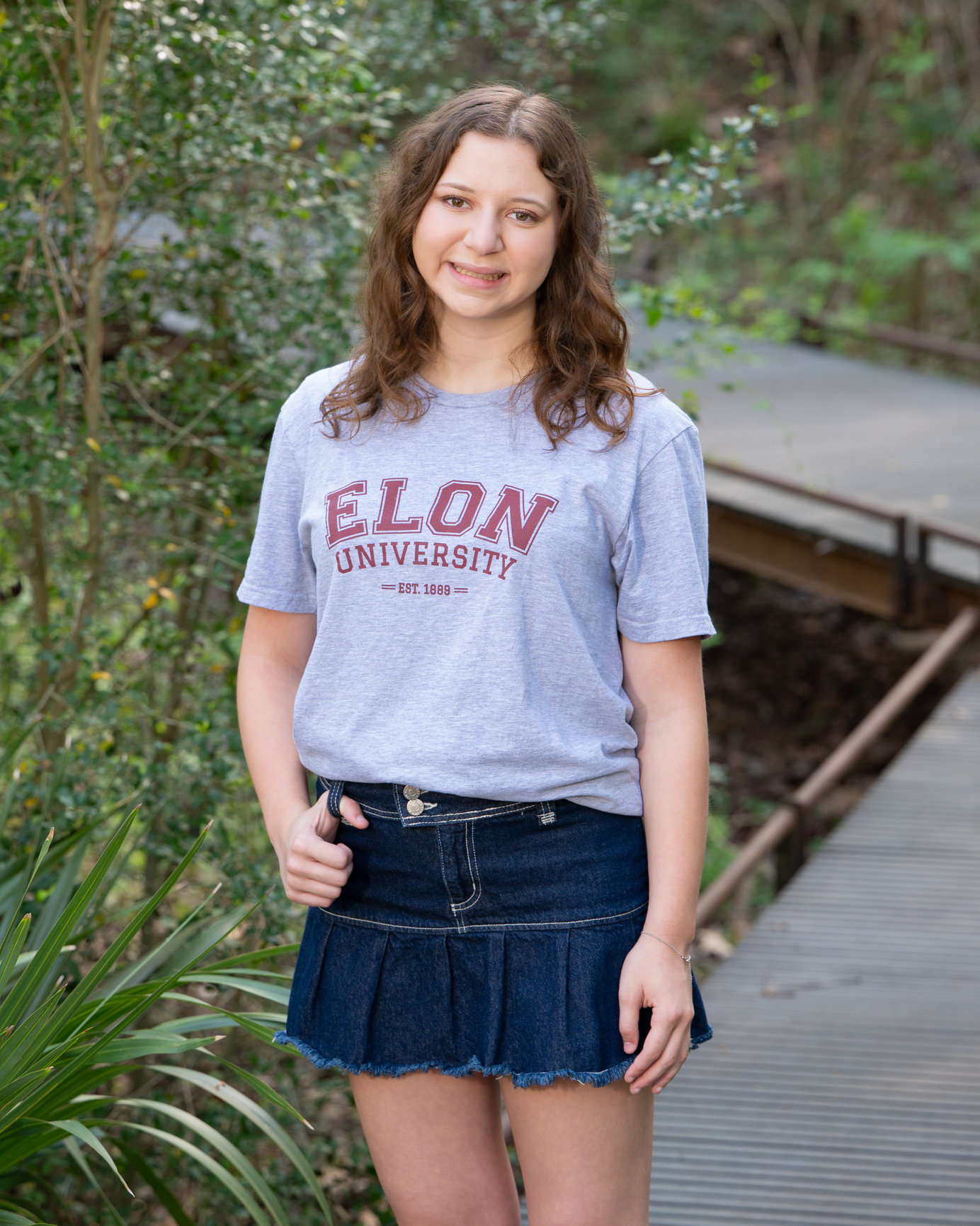 A young woman stands in front of a greenery background with an Elon University t-shirt on