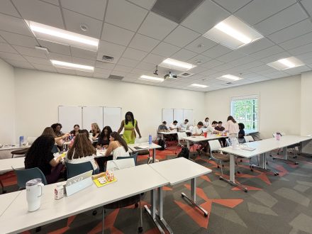 Students work together in small groups around tables in a bright classroom, talking and completing activities with materials spread across their workspaces.