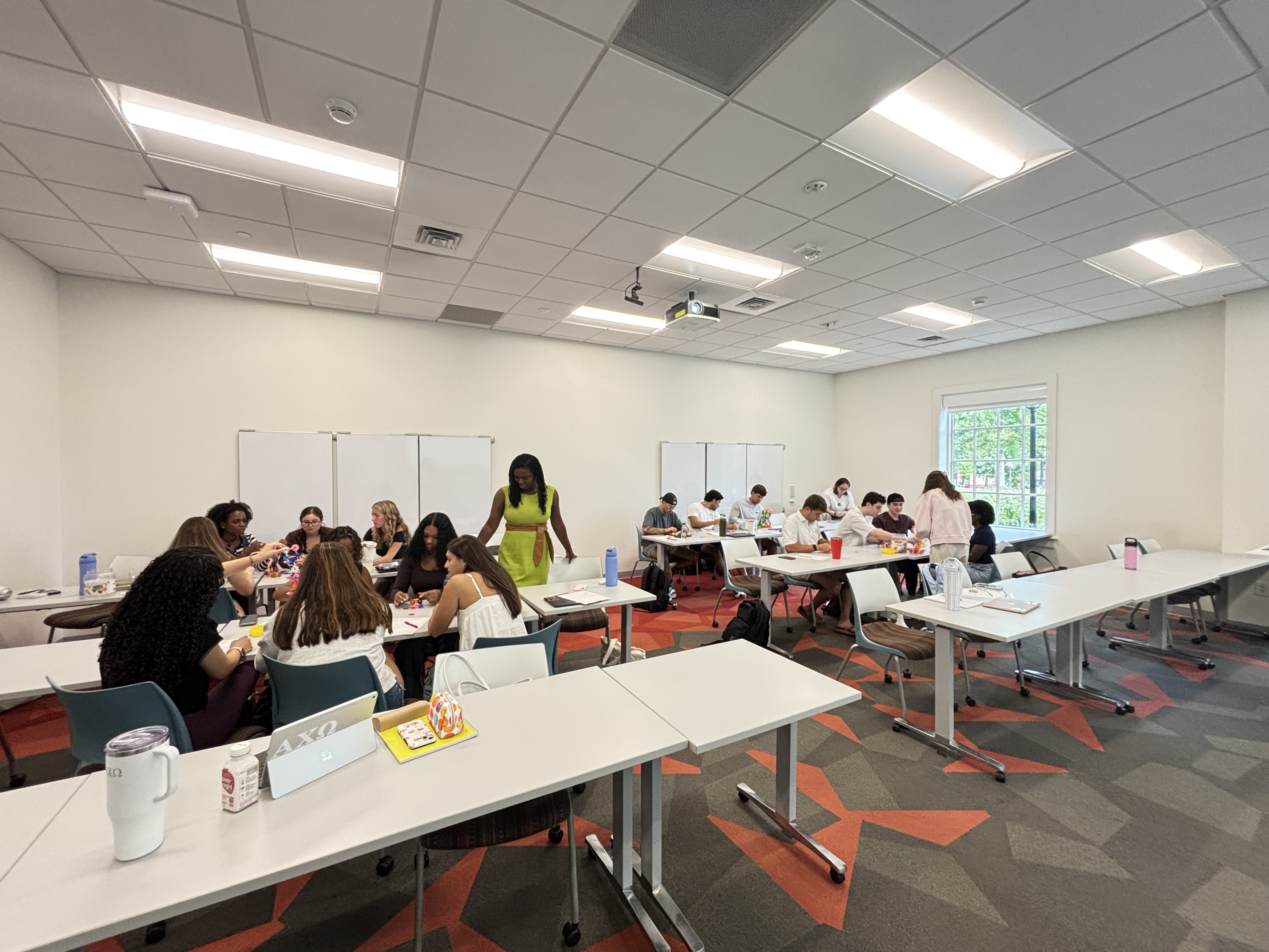 Students work together in small groups around tables in a bright classroom, talking and completing activities with materials spread across their workspaces.