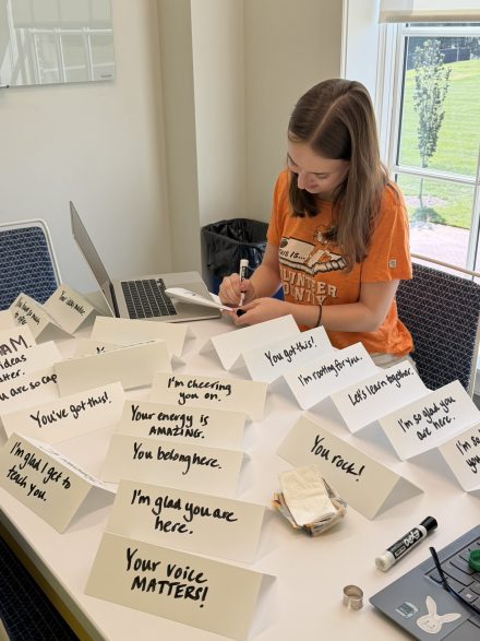Anna Grace Gilbert '27 writing encouraging messages like 'You belong here' and 'You've got this!' on folded table tent cards.