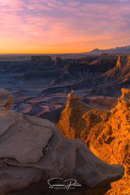 A person stands atop a rocky overlook above a vast desert canyon illuminated by warm sunset light beneath a pink and orange sky.
