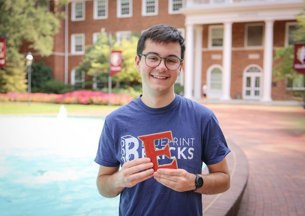 A smiling student wearing glasses holds a red LEGO letter “E” while standing in front of a red-brick campus building.