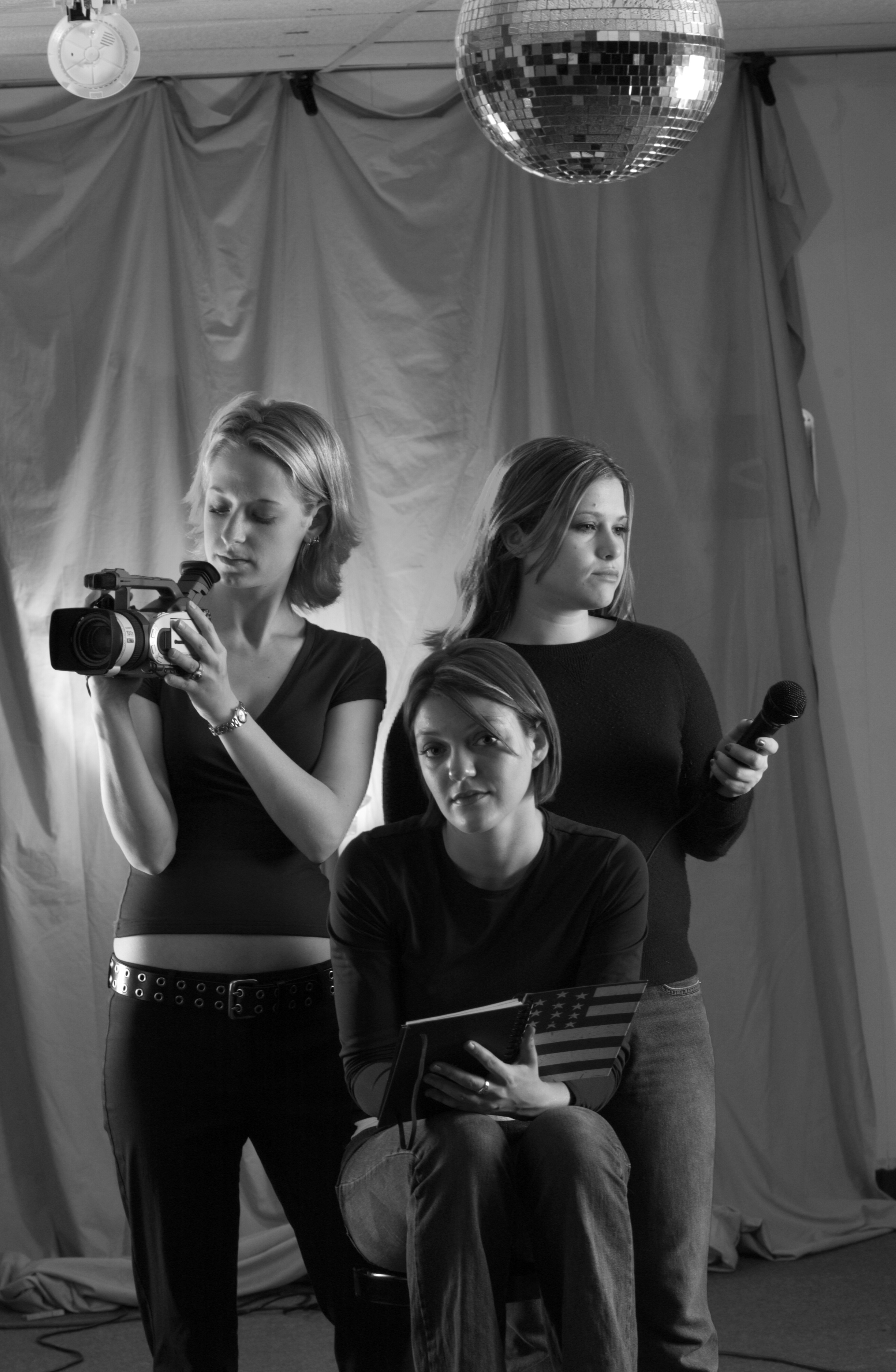 Three students pose with video production equipment, including a video camera, microphone and American flag notebook.