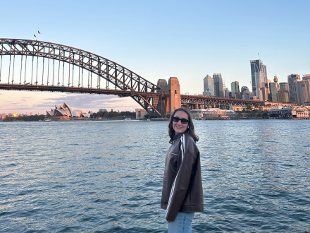 Maria Cassereau smiling by the water with the Sydney Harbour Bridge, Opera House, and city skyline behind her at sunset.