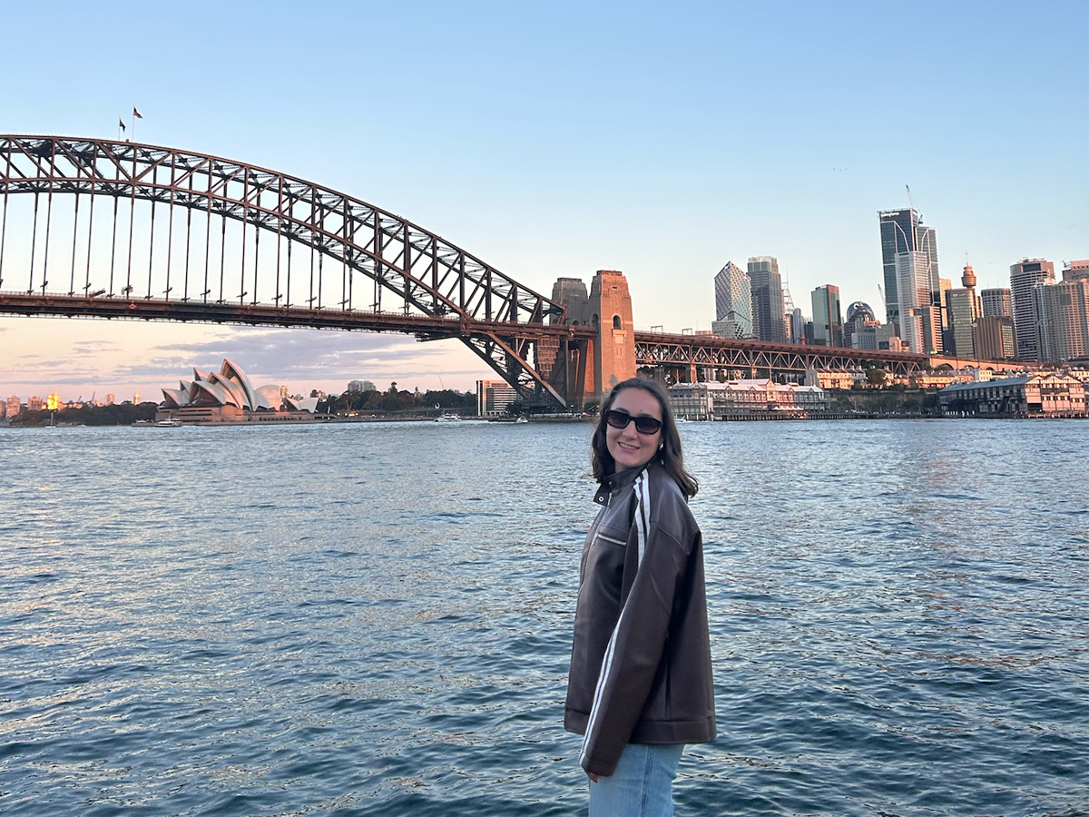 Maria Cassereau smiling by the water with the Sydney Harbour Bridge, Opera House, and city skyline behind her at sunset.
