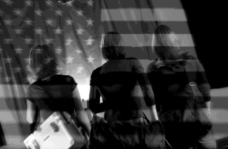Three students carrying equipment walk away from the camera beneath a layered image of an American flag.