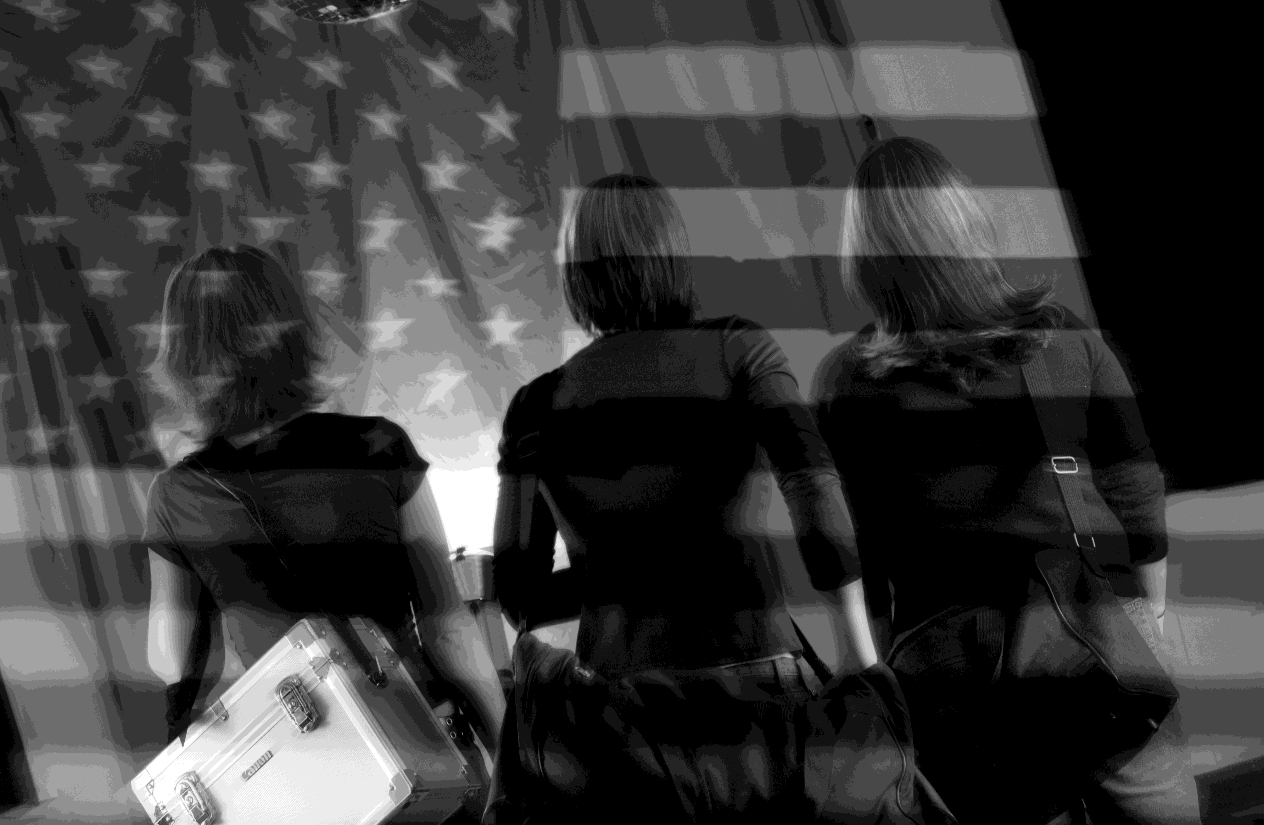 Three students carrying equipment walk away from the camera beneath a layered image of an American flag.
