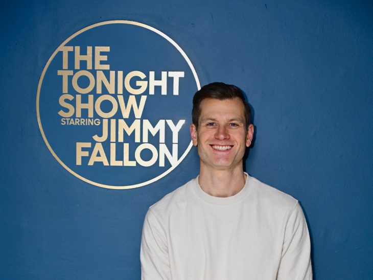Nick Dyer ’11, wearing a white crewneck shirt, smiles beside “The Tonight Show Starring Jimmy Fallon” logo on a blue wall.