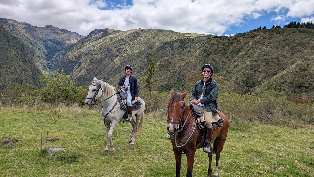 Two people ride horses through a grassy mountain landscape.
