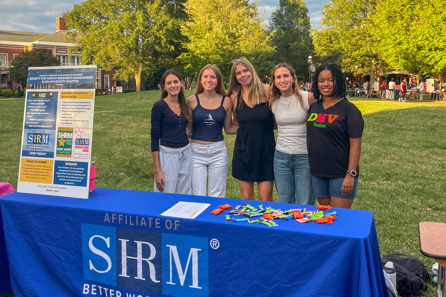 Five Elon students stand behind a Society for Human Resource Management table outdoors on campus. A large SHRM banner and information board are displayed beside them.