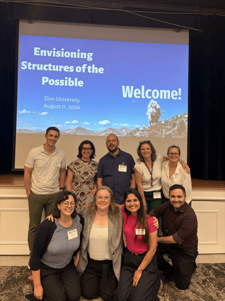 Nine people pose together in two rows at an Elon University event, with an “Envisioning Structures of the Possible” welcome slide behind them.