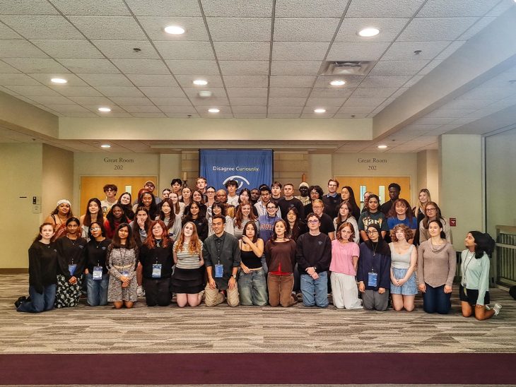 A large group of people pose for a photo in a conference room. A blue banner behind them reads "Disagree Curiously"