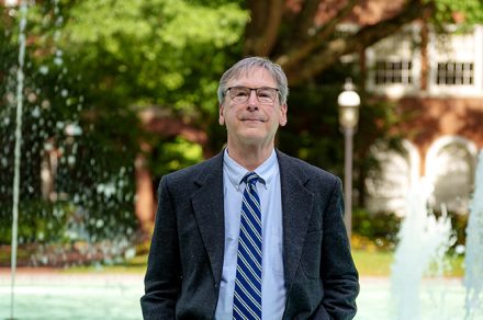 A man in a suit and tie stands knee-deep in an �������޹�����Ʒ fountain, with trees and a brick building in the background.