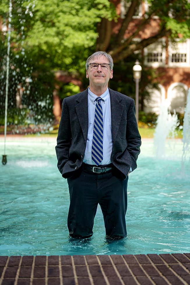 A man in a suit and tie stands knee-deep in an �������޹�����Ʒ fountain, with trees and a brick building in the background.