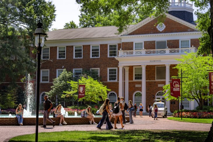 Students walk and gather near Fonville Fountain in front of Alamance Building on a sunny day at Elon University.