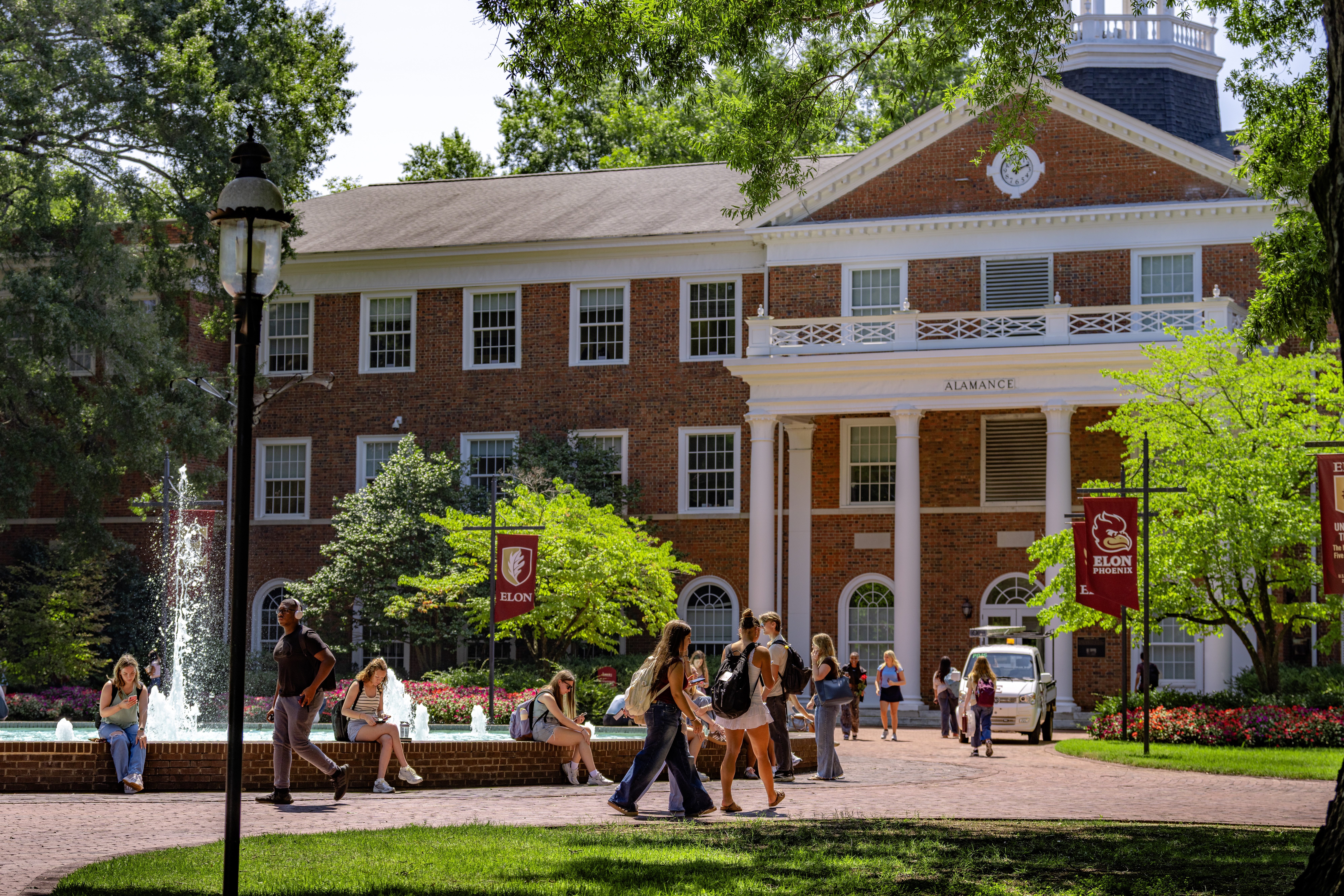 Students walk and gather near Fonville Fountain in front of Alamance Building on a sunny day at Elon University.