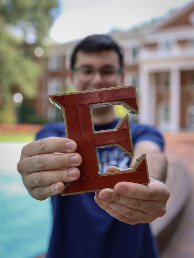 A person holds a red LEGO letter “E” toward the camera, with a red-brick building and white columns blurred in the background.