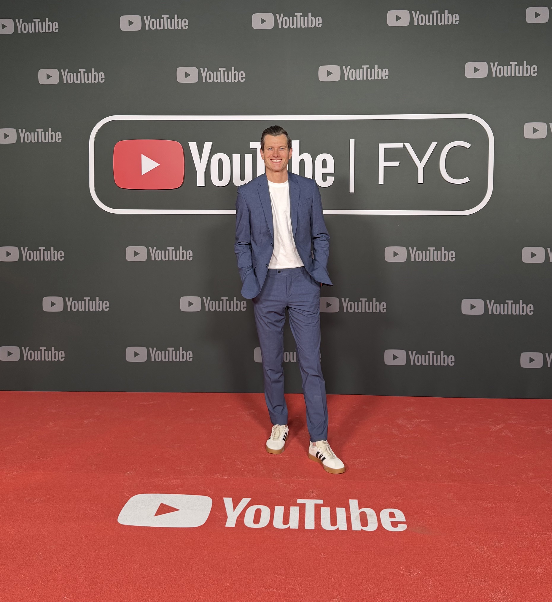 Nick Dyer ’11, wearing a blue suit and white sneakers, stands on a red carpet in front of a YouTube FYC backdrop.