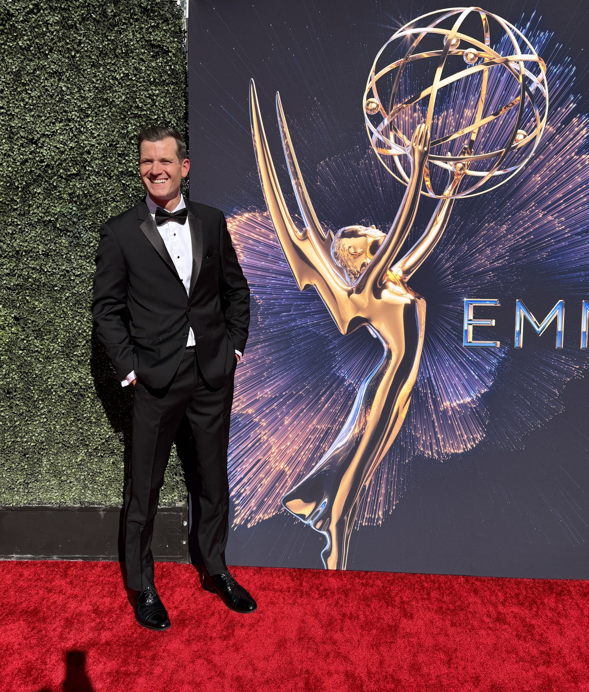 Nick Dyer ’11, wearing a black tuxedo and bow tie, smiles on a red carpet beside a large golden Emmy statuette graphic.